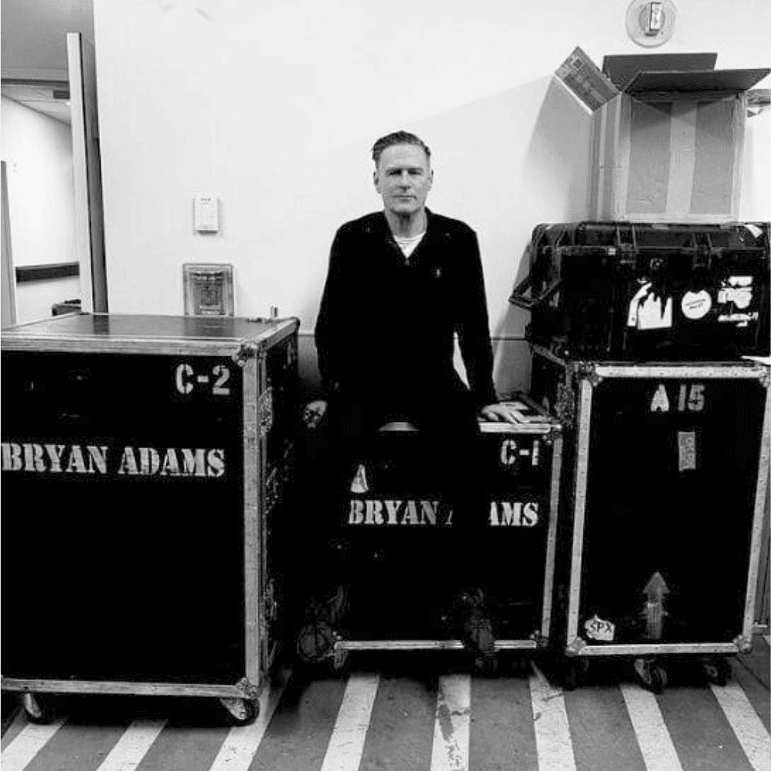 A man in dark clothing sits between large equipment cases labeled BRYAN ADAMS in a backstage area with striped flooring and white walls. The black and white image was taken before his hot 1027 breakfast show performance.