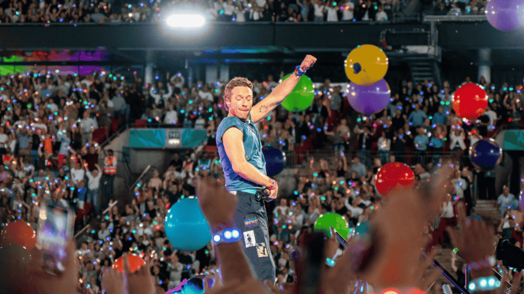 A musician performs energetically on stage in front of a large, cheering crowd at the Hot 1027 Breakfast, with colorful balloons floating and audience members raising their hands and phones.