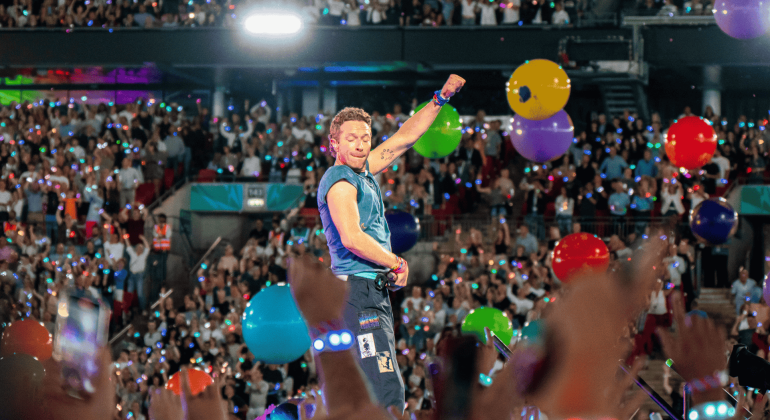 A musician performs energetically on stage in front of a large, cheering crowd at the Hot 1027 Breakfast, with colorful balloons floating and audience members raising their hands and phones.