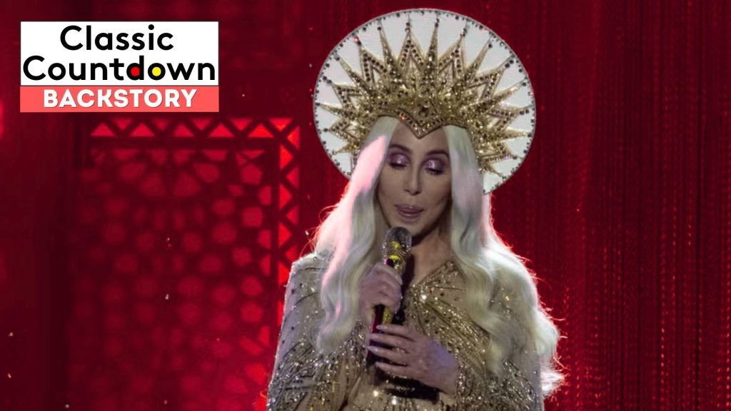 A person with long platinum-blonde hair, wearing a gold, ornate crown and sparkly gold outfit, sings "Last Christmas" into a microphone on stage with a red background. The text Classic Countdown Backstory is in the upper left corner.