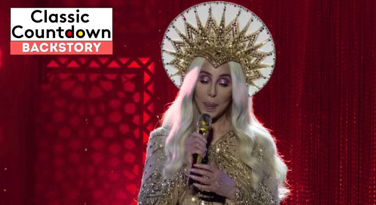 A person with long platinum-blonde hair, wearing a gold, ornate crown and sparkly gold outfit, sings "Last Christmas" into a microphone on stage with a red background. The text Classic Countdown Backstory is in the upper left corner.