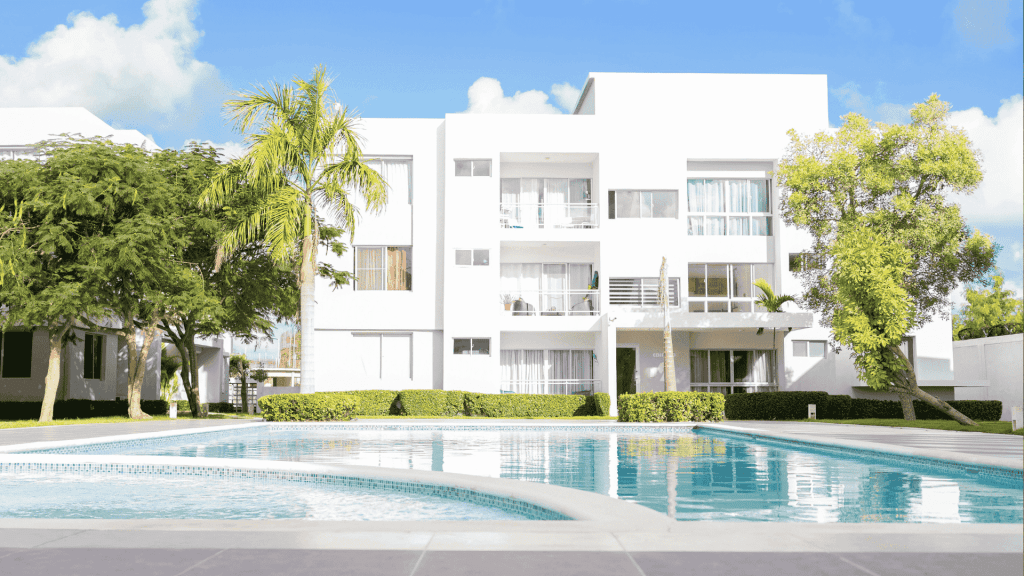 Modern white apartment building with large windows and balconies, surrounded by green trees and bushes, with a clear blue swimming pool in the foreground—perfect for enjoying your hot 1027 breakfast under a bright, sunny sky.