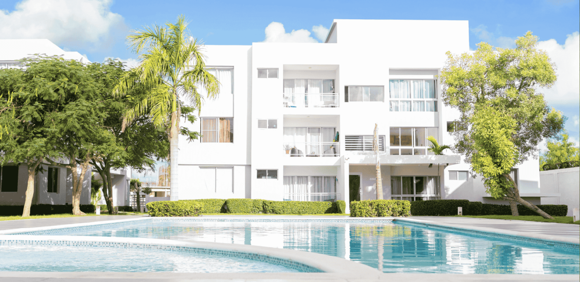 Modern white apartment building with large windows and balconies, surrounded by green trees and bushes, with a clear blue swimming pool in the foreground—perfect for enjoying your hot 1027 breakfast under a bright, sunny sky.