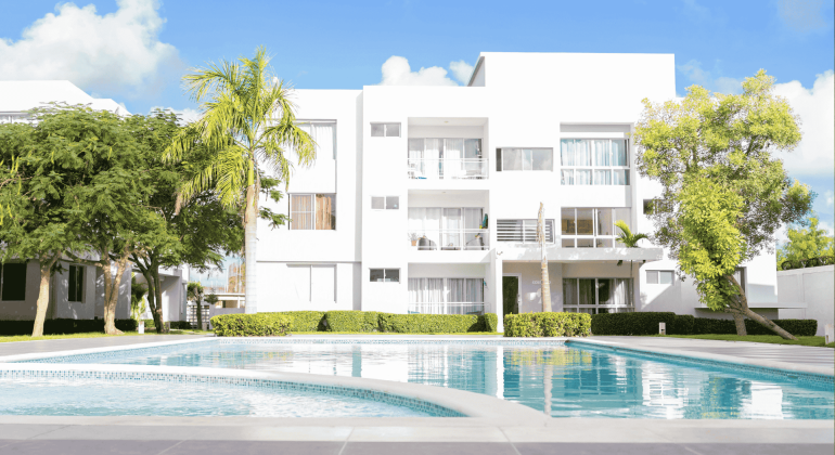 Modern white apartment building with large windows and balconies, surrounded by green trees and bushes, with a clear blue swimming pool in the foreground—perfect for enjoying your hot 1027 breakfast under a bright, sunny sky.