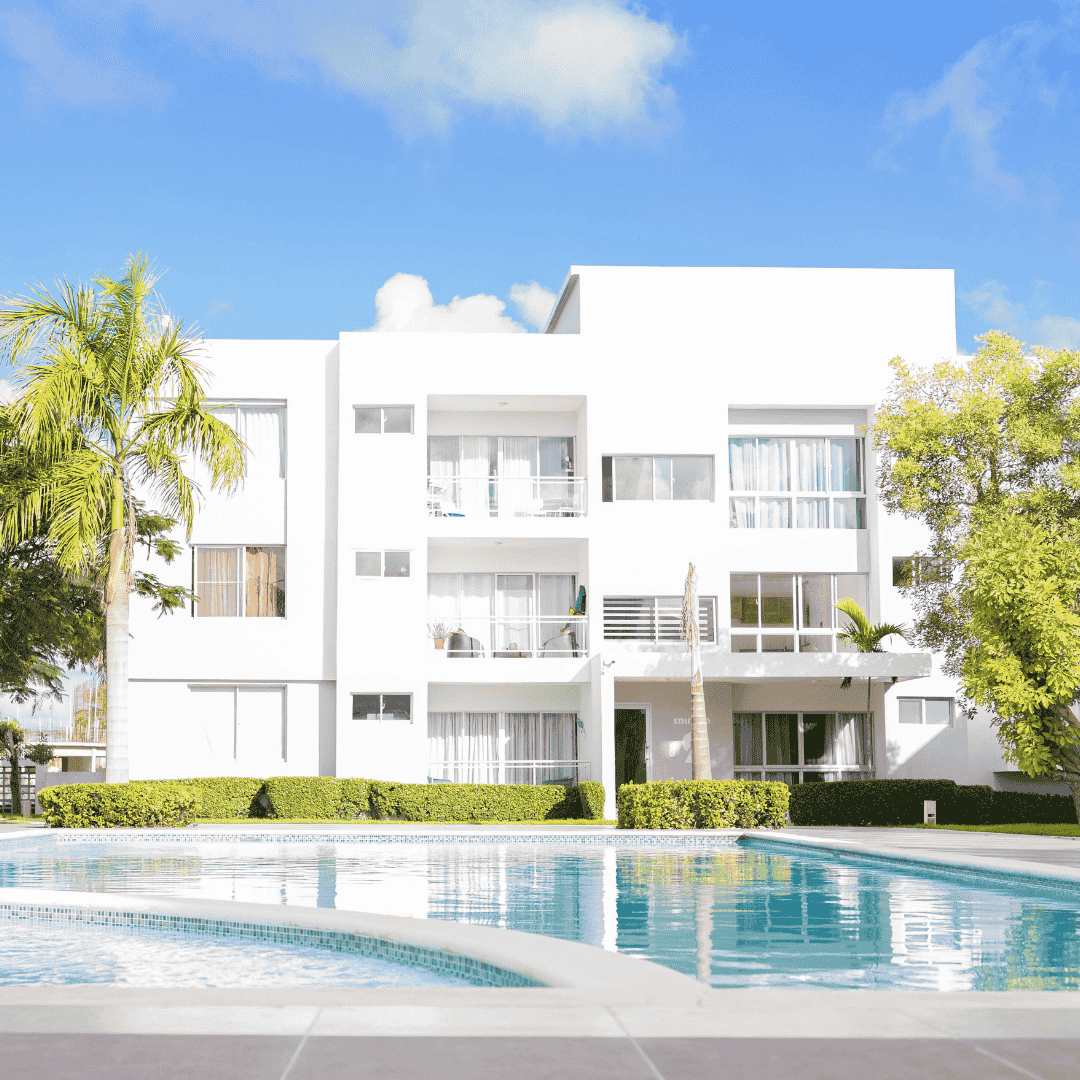 A modern white apartment building with large windows and balconies is surrounded by palm trees and greenery, with a clear blue swimming pool in the foreground—perfect for enjoying a hot 1027 breakfast under a bright, sunny sky.