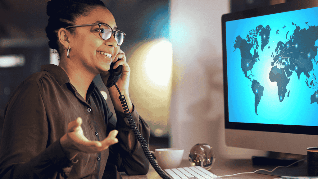 A woman smiles while talking on the phone at her desk. She gestures with her hand, facing a computer screen displaying Matric’s world map interface, with connection lines linking different countries.