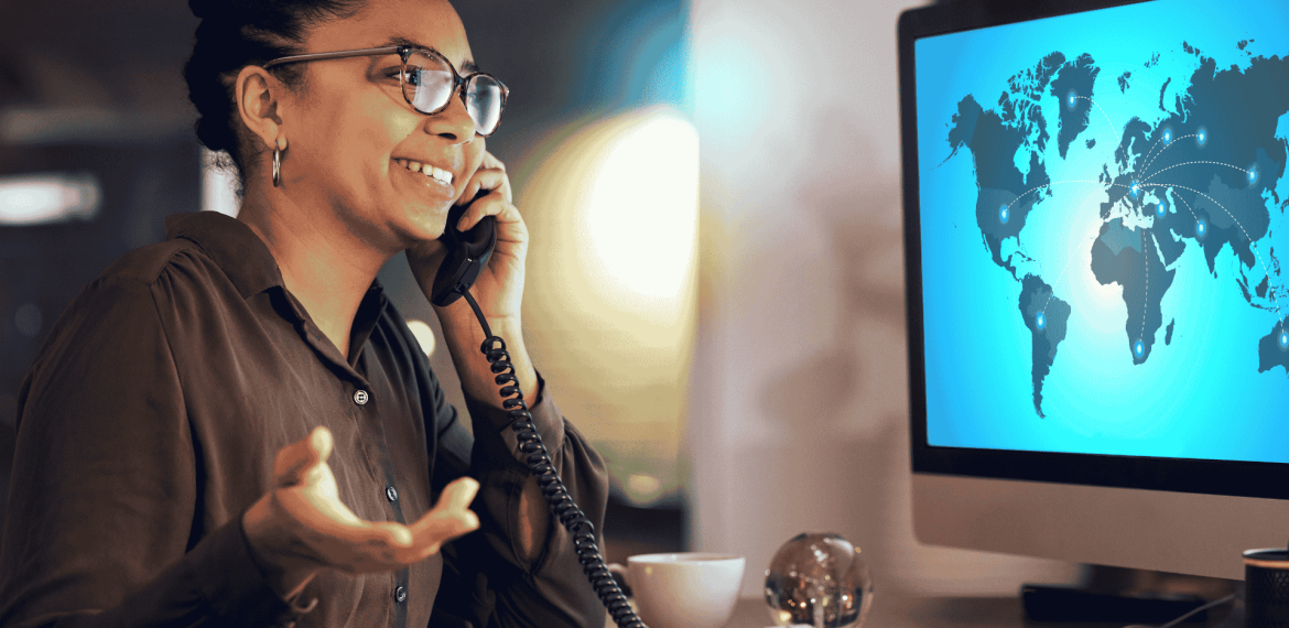 A woman smiles while talking on the phone at her desk. She gestures with her hand, facing a computer screen displaying Matric’s world map interface, with connection lines linking different countries.
