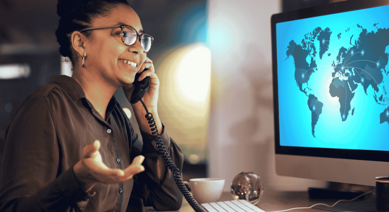 A woman smiles while talking on the phone at her desk. She gestures with her hand, facing a computer screen displaying Matric’s world map interface, with connection lines linking different countries.