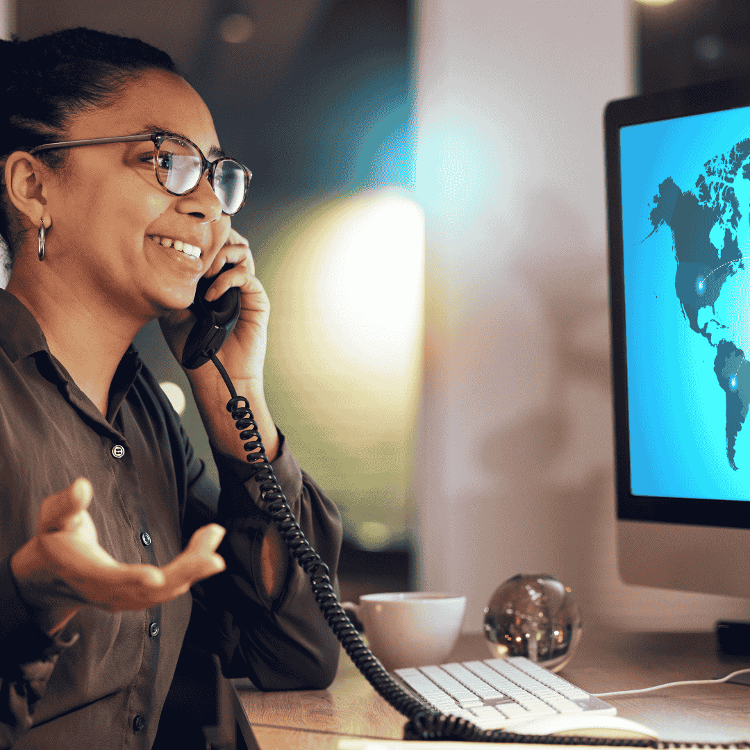 A person wearing glasses smiles while talking on a landline phone at a desk, gesturing with one hand. A computer monitor shows a world map, while a coffee cup, globe, and Matric documents are also on the desk.