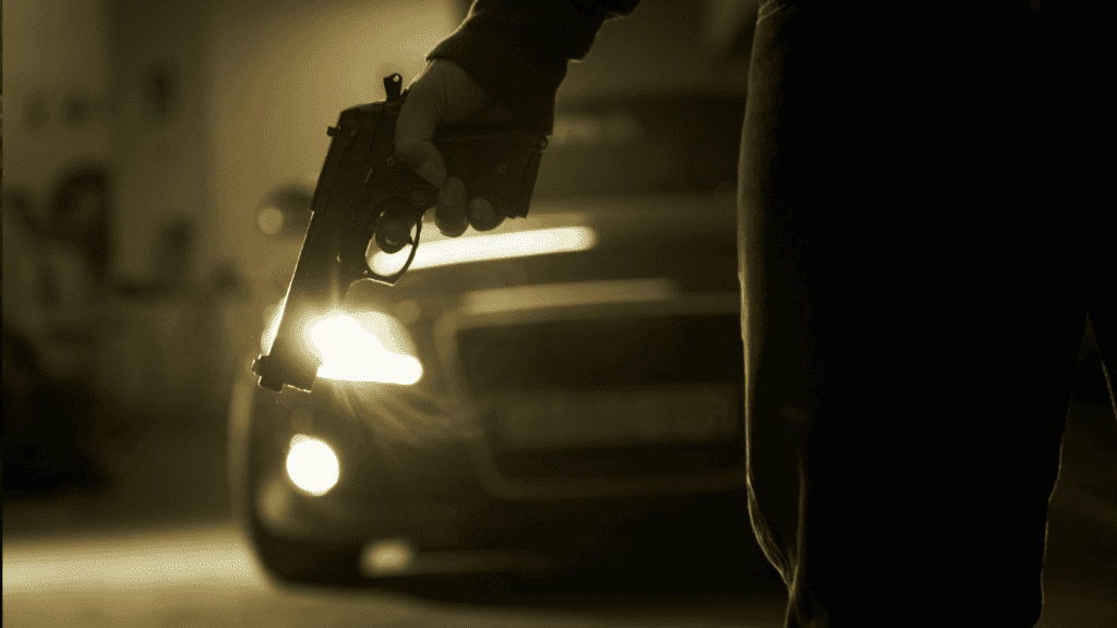 A person holds a handgun down by their side in a dimly lit area, with a car and its headlights creating a dramatic, tense mood—like the suspense before the hot 1027 breakfast show kicks off the morning.