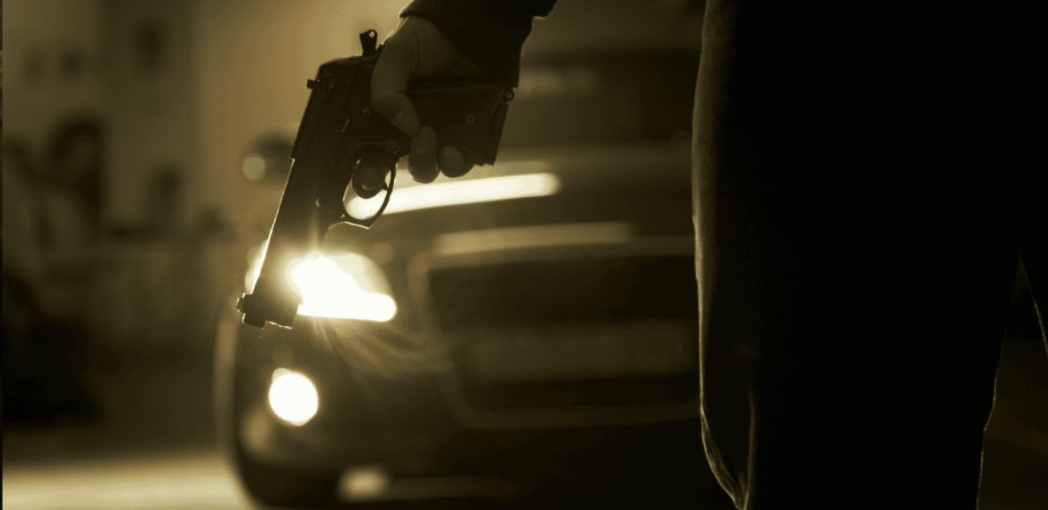 A person holds a handgun down by their side in a dimly lit area, with a car and its headlights creating a dramatic, tense mood—like the suspense before the hot 1027 breakfast show kicks off the morning.