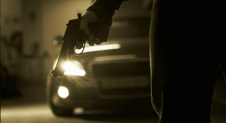 A person holds a handgun down by their side in a dimly lit area, with a car and its headlights creating a dramatic, tense mood—like the suspense before the hot 1027 breakfast show kicks off the morning.