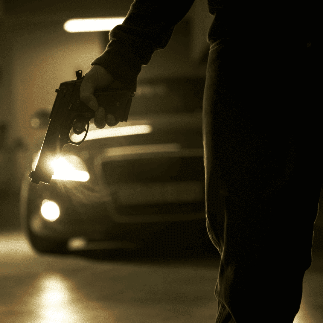 A person in dark clothing holds a handgun in a dimly lit area near a car with headlights on, evoking the tense suspense of a late-night drama—far from your typical hot 1027 breakfast scene.