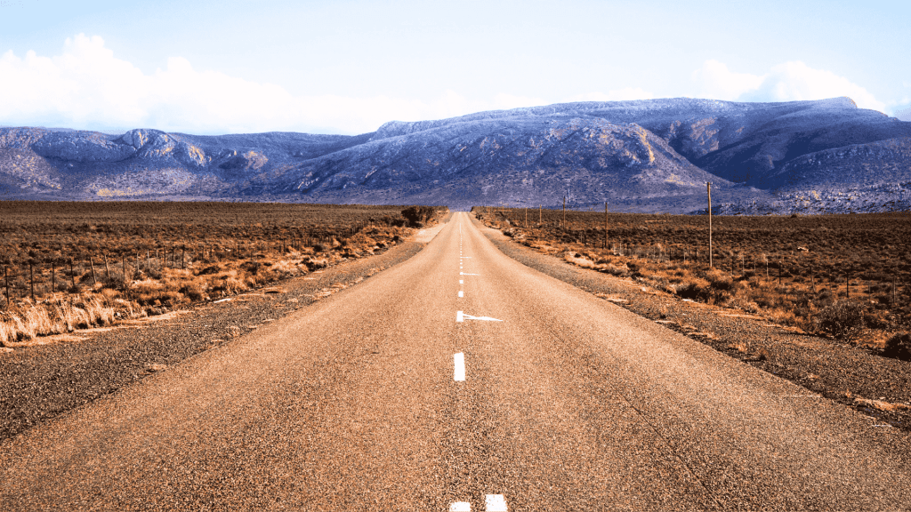 A straight, empty road stretches into the distance through a dry, brown landscape with mountains in the background under a partly cloudy sky, evoking the feeling of setting out on a new journey after completing matric.