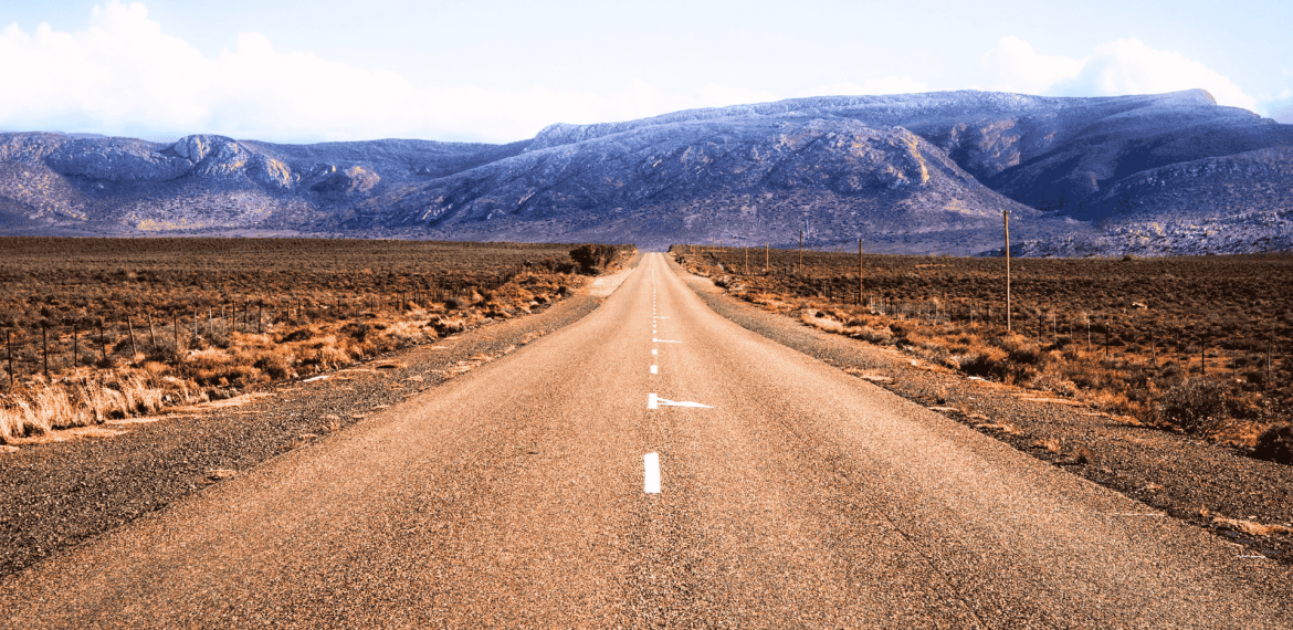 A straight, empty road stretches into the distance through a dry, brown landscape with mountains in the background under a partly cloudy sky, evoking the feeling of setting out on a new journey after completing matric.