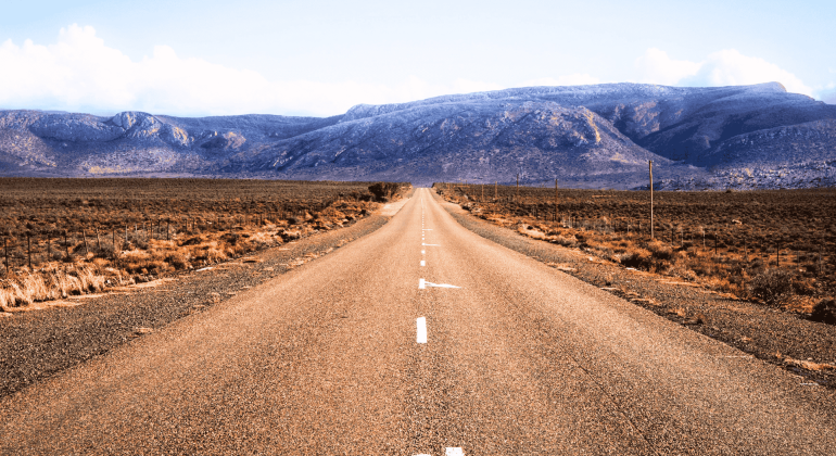A straight, empty road stretches into the distance through a dry, brown landscape with mountains in the background under a partly cloudy sky, evoking the feeling of setting out on a new journey after completing matric.