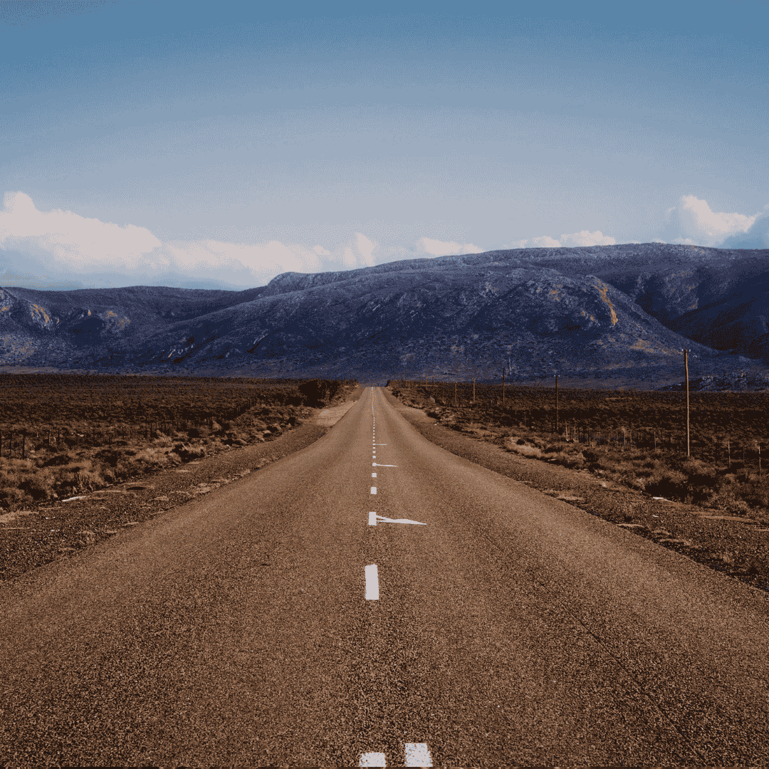 A straight, empty road stretches into the distance through a dry, flat landscape—its path almost like a matric grid—with mountains in the background under a blue sky with scattered clouds.