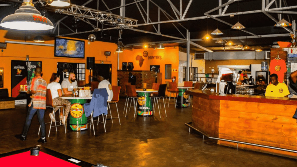 A brightly lit bar with orange walls, scattered tables and chairs, people socializing over hot 1027 breakfast, two TV screens, a pool table in the foreground, and a wooden counter on the right.