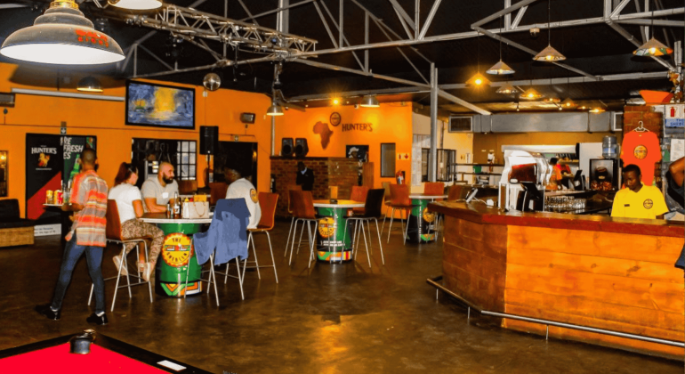 A brightly lit bar with orange walls, scattered tables and chairs, people socializing over hot 1027 breakfast, two TV screens, a pool table in the foreground, and a wooden counter on the right.