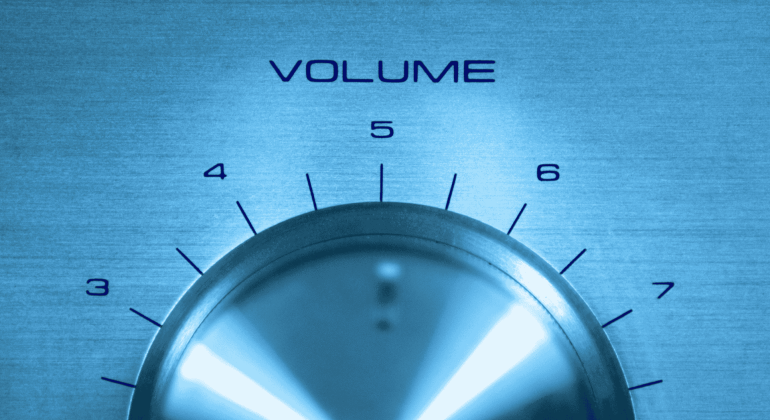 A close-up of a metallic volume control knob set between levels 5 and 6, with numbers 4 to 7 visible above the knob and "VOLUME" printed above in blue text. The blue-tinted image channels the early energy of a hot 1027 breakfast show.