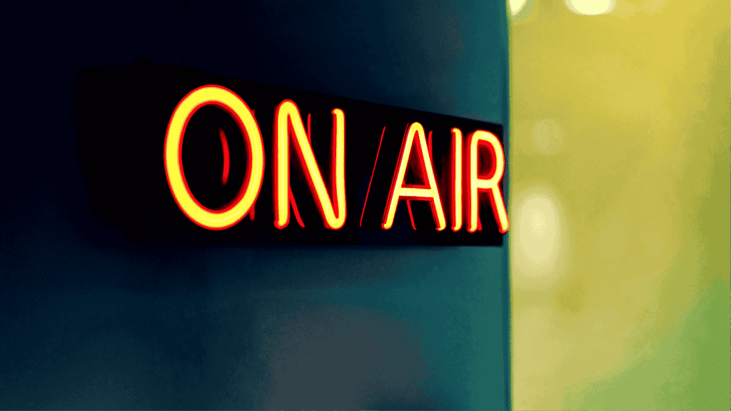 A bright neon sign reading ON AIR glows in red and yellow against a dark, blurred background, signaling that the Hot 1027 Breakfast live broadcast or recording is currently in progress.