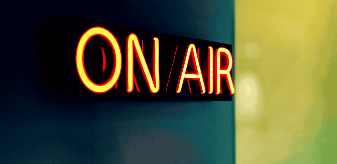 A bright neon sign reading ON AIR glows in red and yellow against a dark, blurred background, signaling that the Hot 1027 Breakfast live broadcast or recording is currently in progress.