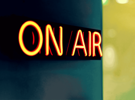 A bright neon sign reading ON AIR glows in red and yellow against a dark, blurred background, signaling that the Hot 1027 Breakfast live broadcast or recording is currently in progress.