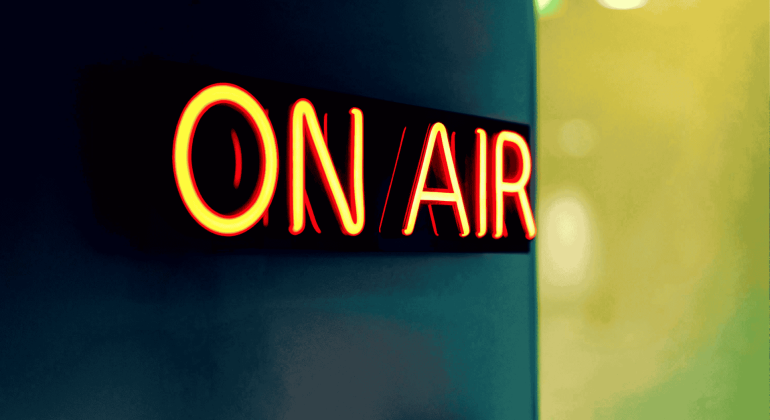 A bright neon sign reading ON AIR glows in red and yellow against a dark, blurred background, signaling that the Hot 1027 Breakfast live broadcast or recording is currently in progress.