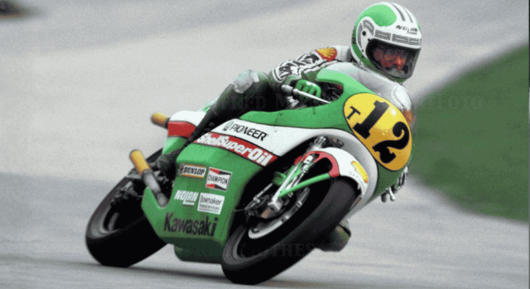 Mark Andrews, a motorcycle racer wearing a green and white suit and helmet, leans into a turn on his green Kawasaki bike numbered 12 during a race on a paved track.