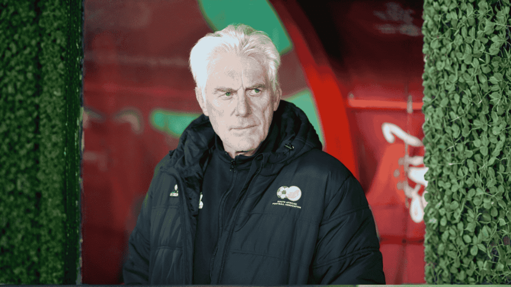 An older man with white hair in a dark jacket stands before red and green colors, leafy green bush walls on either side. With a serious expression, he looks slightly to the side, as if contemplating his next travel getaway.