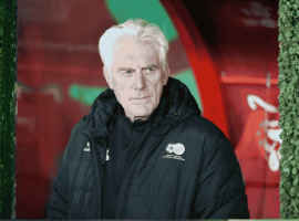 An older man with white hair in a dark jacket stands before red and green colors, leafy green bush walls on either side. With a serious expression, he looks slightly to the side, as if contemplating his next travel getaway.