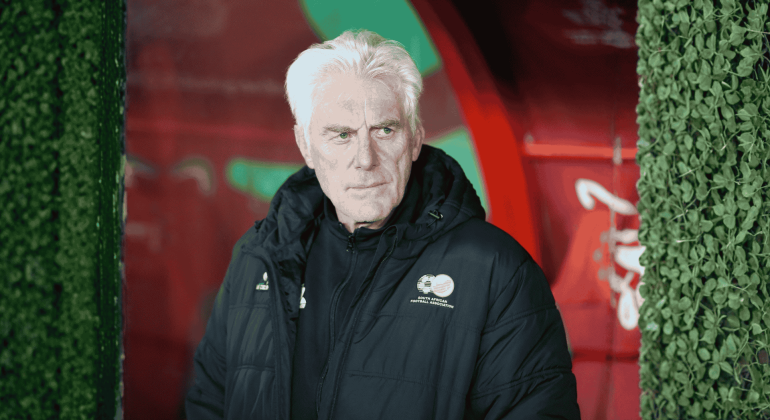 An older man with white hair in a dark jacket stands before red and green colors, leafy green bush walls on either side. With a serious expression, he looks slightly to the side, as if contemplating his next travel getaway.