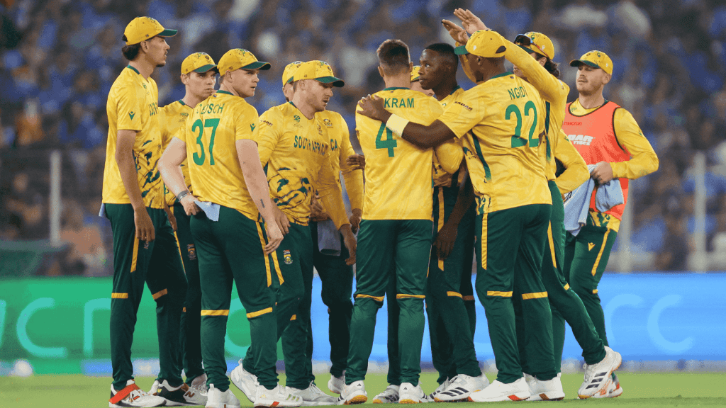 A group of South African cricket players in yellow and green uniforms huddle together on the field during a match, with a crowd visible in the blurred background, capturing the spirit much like a lively Hot 1027 Breakfast gathering.