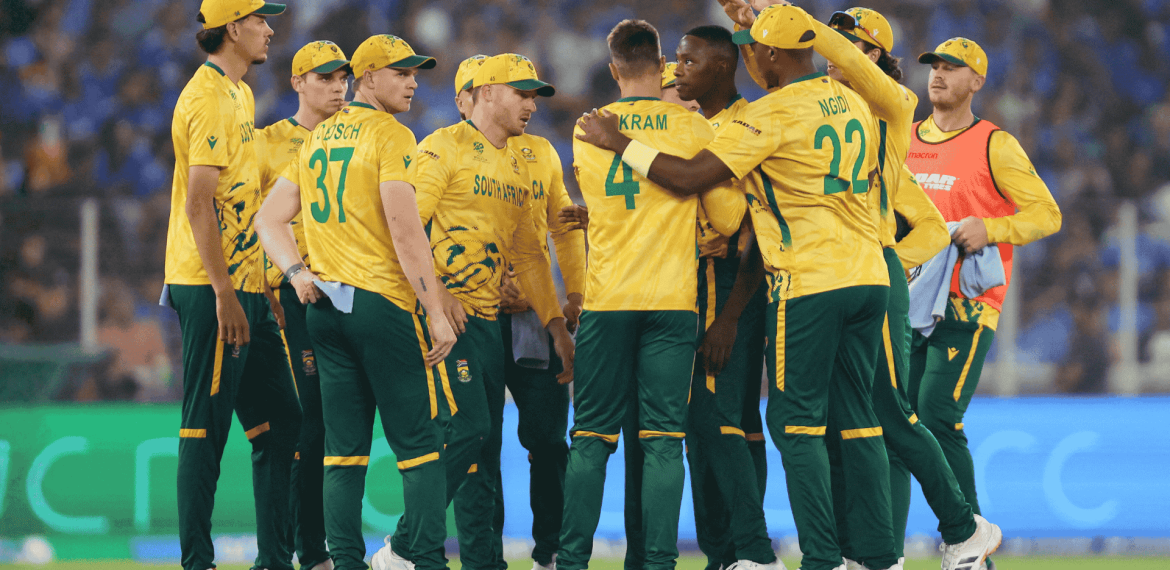 A group of South African cricket players in yellow and green uniforms huddle together on the field during a match, with a crowd visible in the blurred background, capturing the spirit much like a lively Hot 1027 Breakfast gathering.
