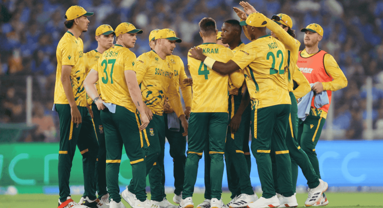 A group of South African cricket players in yellow and green uniforms huddle together on the field during a match, with a crowd visible in the blurred background, capturing the spirit much like a lively Hot 1027 Breakfast gathering.