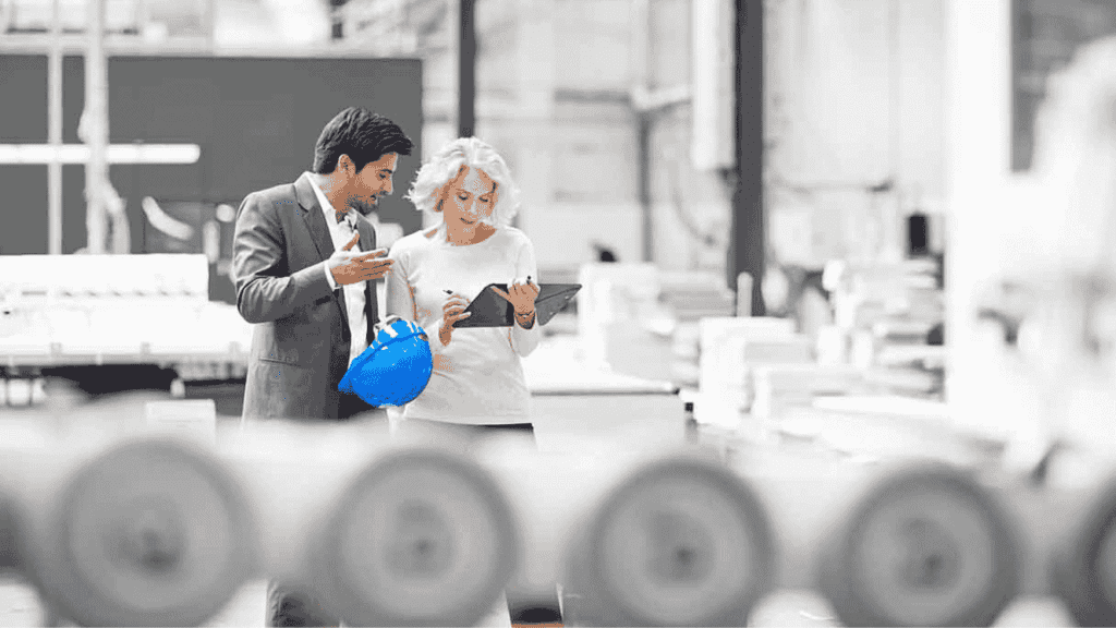 Two professionals, one holding a blue hard hat, stand in a bright industrial setting. As they discuss plans over a tablet, the energy feels as lively as a Hot 1027 Breakfast, with machinery and blurred equipment in the foreground.