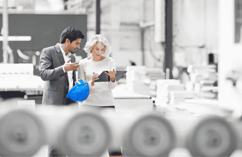 Two professionals, one holding a blue hard hat, stand in a bright industrial setting. As they discuss plans over a tablet, the energy feels as lively as a Hot 1027 Breakfast, with machinery and blurred equipment in the foreground.