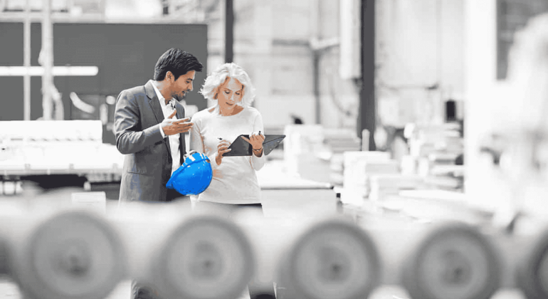 Two professionals, one holding a blue hard hat, stand in a bright industrial setting. As they discuss plans over a tablet, the energy feels as lively as a Hot 1027 Breakfast, with machinery and blurred equipment in the foreground.