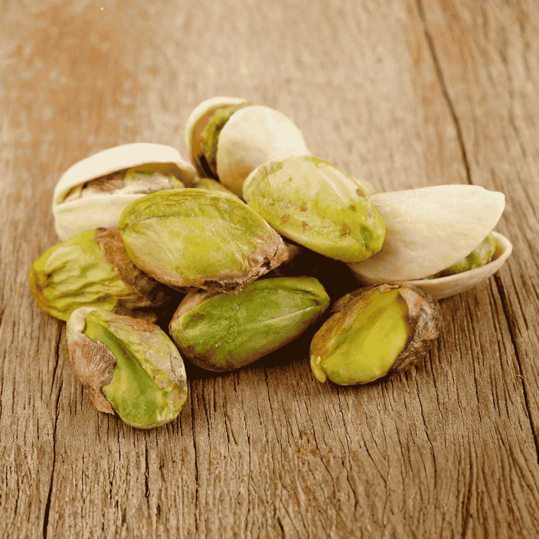 A pile of unshelled and shelled pistachio nuts on a rustic wooden surface, creating a natural matric of textures. The green nuts are partially exposed, with empty and whole shells mixed together.