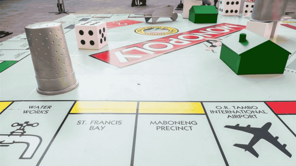 A large Monopoly board with oversized game pieces, including a thimble, dice, and houses, features properties like Water Works, St. Francis Bay, Maboneng Precinct, O.R. Tambo International Airport—and even the excitement of Hot 1027 Breakfast.