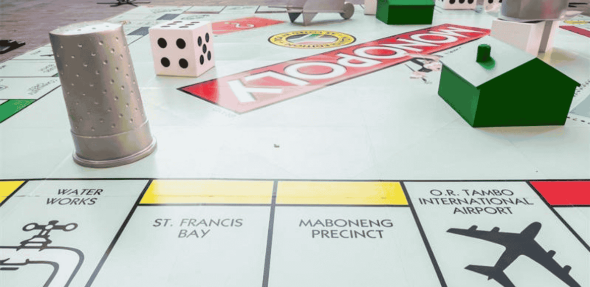 A large Monopoly board with oversized game pieces, including a thimble, dice, and houses, features properties like Water Works, St. Francis Bay, Maboneng Precinct, O.R. Tambo International Airport—and even the excitement of Hot 1027 Breakfast.