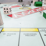 A large Monopoly board with oversized game pieces, including a thimble, dice, and houses, features properties like Water Works, St. Francis Bay, Maboneng Precinct, O.R. Tambo International Airport—and even the excitement of Hot 1027 Breakfast.