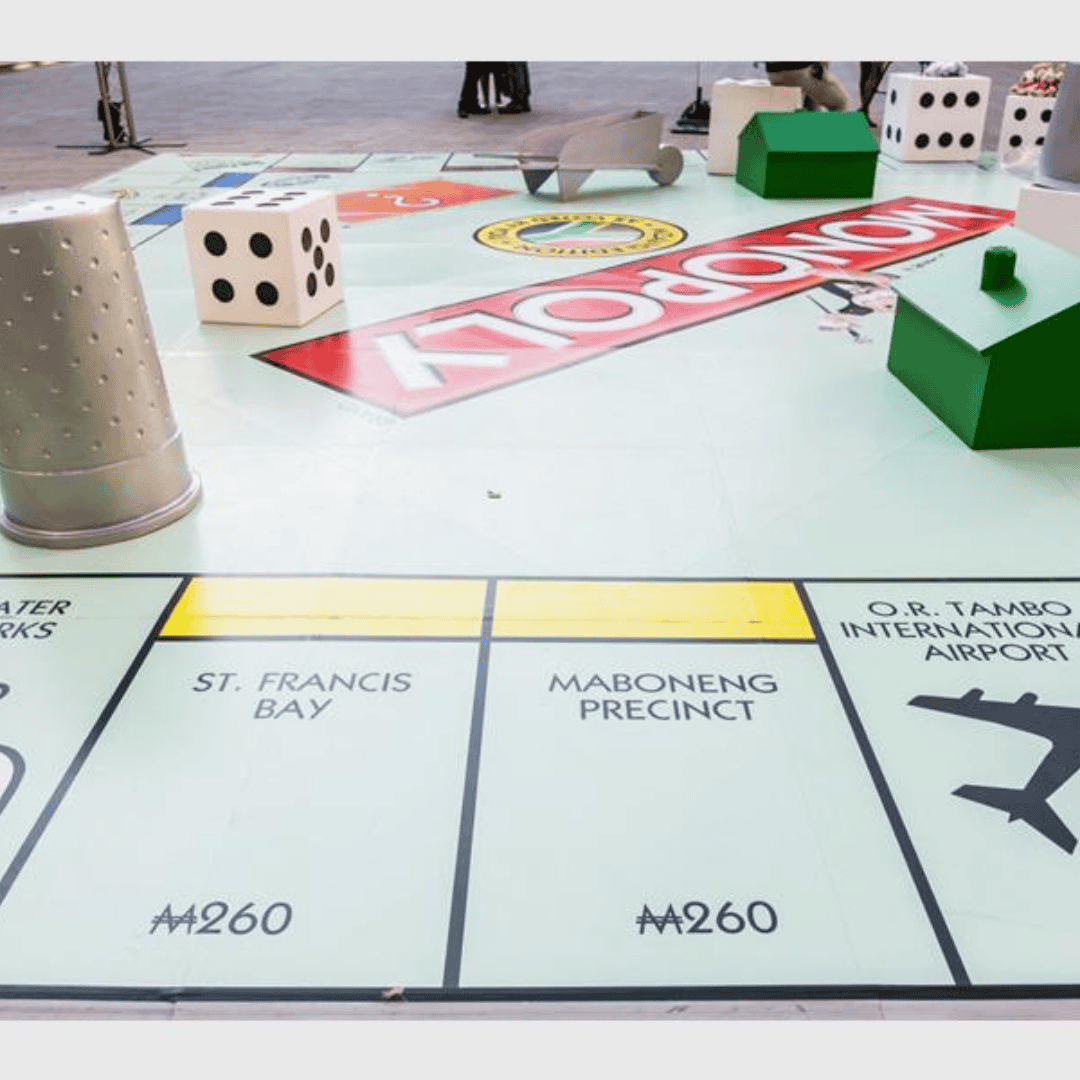 A giant Monopoly board is set up outdoors, featuring oversized dice, houses, and game pieces. Enjoy fun with locations like St. Francis Bay and O.R. Tambo International Airport—perfect for a Hot 1027 Breakfast event!.