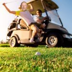 Two people are near a golf cart on a sunny golf course. One person is swinging a golf club, smiling, while the other watches from the driver’s seat—enjoying their morning with Hot 1027 Breakfast as a golf ball sits on the grass in the foreground.