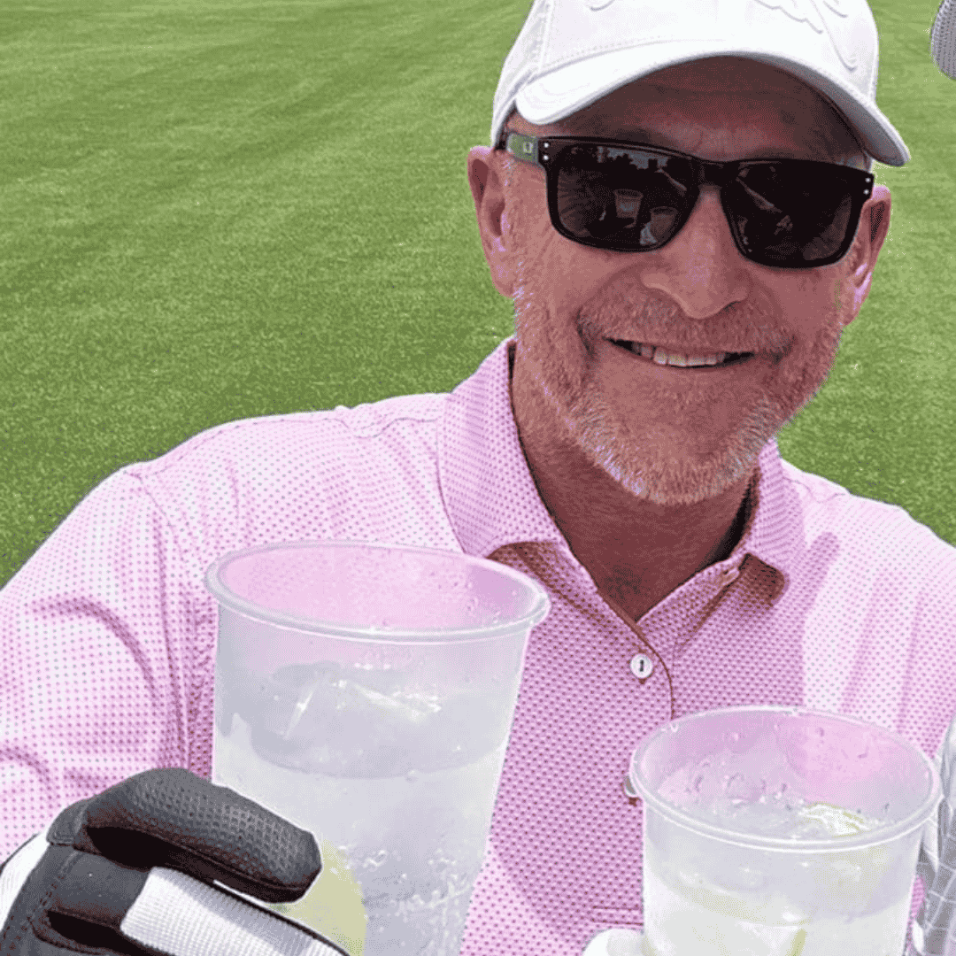 A smiling man in sunglasses, a white cap, and a pink polo shirt holds up a plastic cup with a drink while standing on a golf course during the Hot 1027 Breakfast event. Another gloved hand with a drink is visible in the foreground.