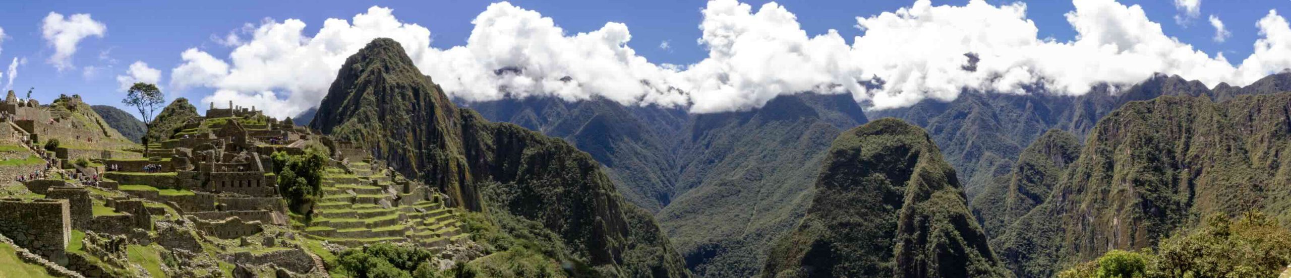 Panoramic view of Machu Picchu’s ancient stone ruins and terraced hills, surrounded by lush green mountains and bush-covered slopes—a breathtaking travel getaway under a blue sky with scattered white clouds.