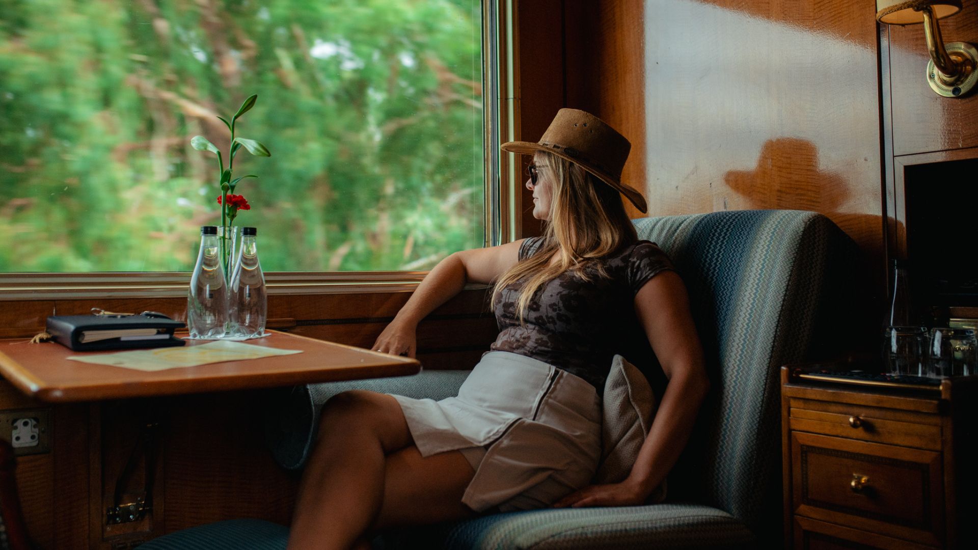 A woman wearing a hat sits on a train seat by a window, gazing out at blurred green bush scenery. The table beside her, set with water bottles, a menu, and a plant, adds comfort to her peaceful travel getaway.