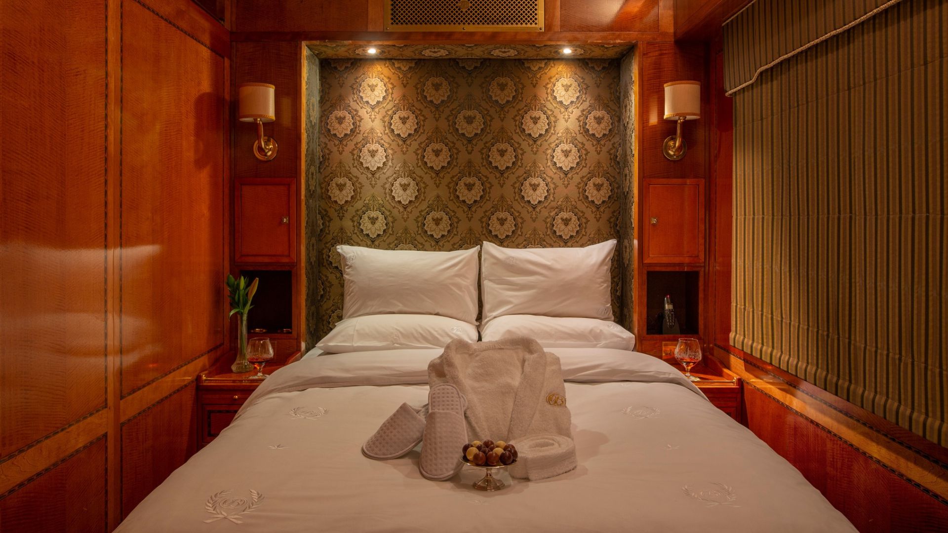 A luxurious bed with white linens, towels, slippers, and robes neatly arranged on top offers the perfect travel getaway in an elegant wood-paneled room with patterned wallpaper, bedside lamps, and wine glasses on nightstands.