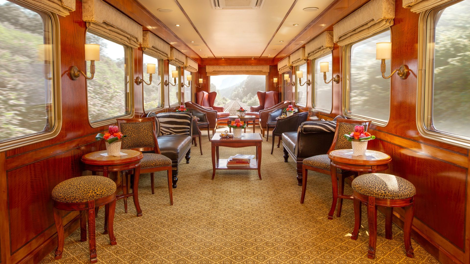 Luxurious train lounge car with polished wood walls, large windows, elegant upholstered chairs, small round tables with flower vases, and soft ambient lighting—perfect for a scenic travel getaway through the bush.