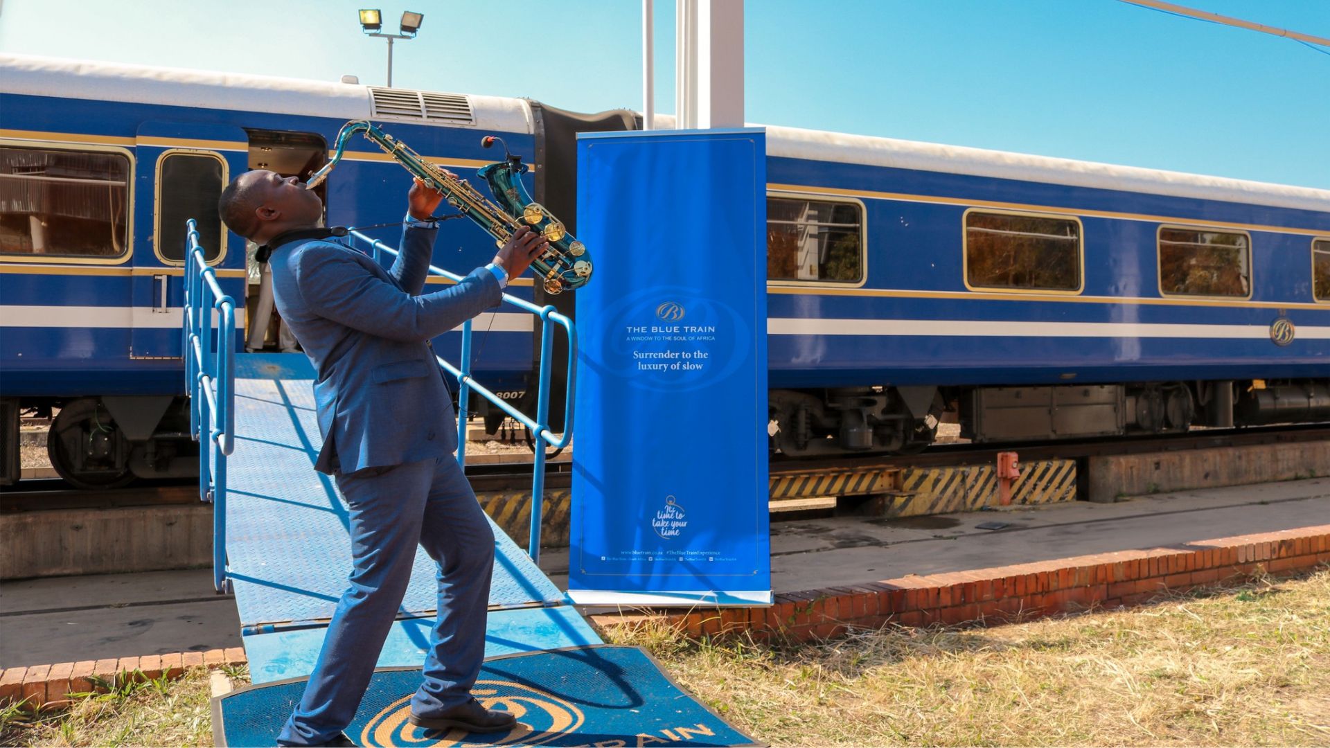 A man in a blue suit plays the saxophone passionately on a blue metal ramp beside a luxury blue train—perfect for a travel getaway—with a blue banner reading, “The Blue Train: Sometimes it's the journey that matters.”.